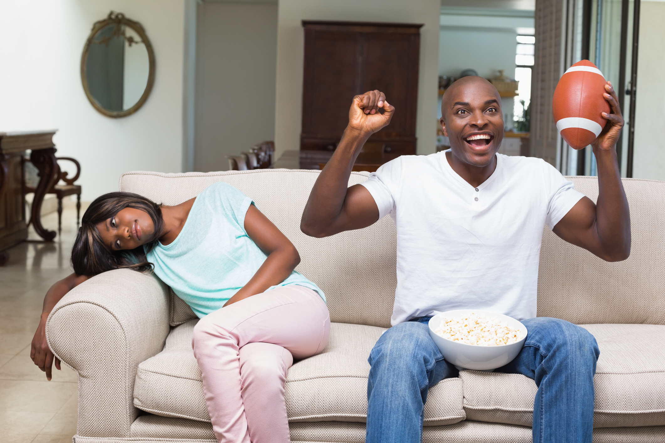Bored woman sitting next to her boyfriend watching football at home in the living room.